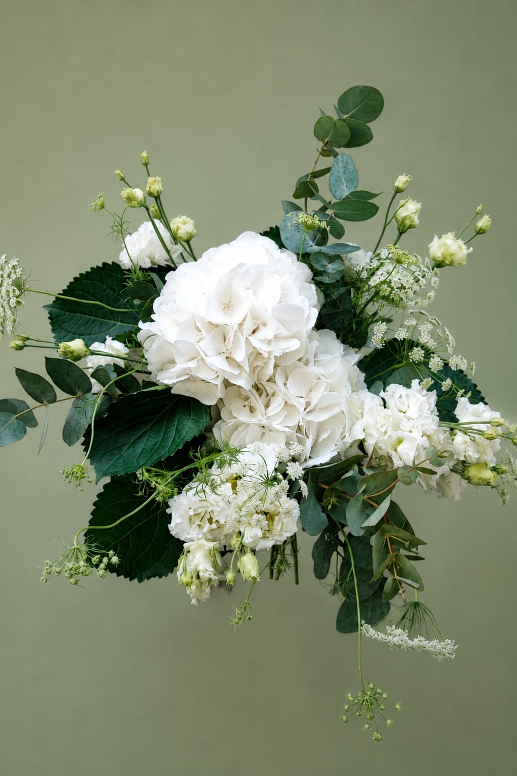 Photo produit du bouquet « Comme au Jardin — Automne » sur fond vert olive uni. La composition florale blanche associe de grands hortensias blancs, de petites fleurs blanches délicates, des feuillages d’eucalyptus vert bleuté et plusieurs branches fines et aériennes. Le bouquet présente une silhouette naturelle, asymétrique et champêtre avec des tiges retombantes. Visuel e-commerce pour commander un bouquet de fleurs et une livraison à Caen. Aucun texte ni prix visible.