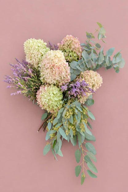 Photo produit du bouquet « Comme au Jardin — Automne » de Rouge Pivoine, posé sur un fond rose poudré. La composition verticale associe de gros hortensias crème, vert tendre et rose pâle, des petites fleurs mauves et un feuillage d’eucalyptus bleu-vert qui s’étire sur la droite et vers le bas. Bouquet de saison au style champêtre et naturel, photographié à plat pour la boutique fleuriste à Caen. Aucun texte, logo ou prix n’est visible.