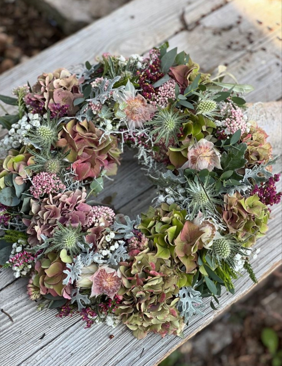 Photo d’inspiration pour l’atelier « Couronne d’Automne 2026 » de Rouge Pivoine à Caen. Couronne florale ronde posée sur une table en bois gris, composée d’hortensias vert olive et vieux rose, chardons bleu-gris, petites fleurs roses et bordeaux, baies, eucalyptus et feuillages variés. La composition présente des tons naturels, poudrés et automnaux avec une texture dense et végétale. Aucun texte, logo ou prix visible.