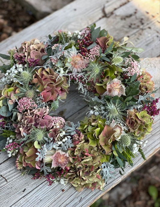Photo d’inspiration pour l’atelier « Couronne d’Automne 2026 » de Rouge Pivoine à Caen. Couronne florale ronde posée sur une table en bois gris, composée d’hortensias vert olive et vieux rose, chardons bleu-gris, petites fleurs roses et bordeaux, baies, eucalyptus et feuillages variés. La composition présente des tons naturels, poudrés et automnaux avec une texture dense et végétale. Aucun texte, logo ou prix visible.