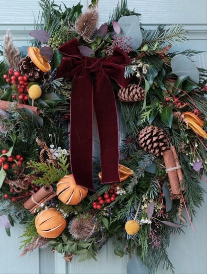 Photo d’inspiration pour l’atelier « Couronne de l’Avent 2026 » de Rouge Pivoine à Caen. Grande couronne de Noël végétale composée de branches de sapin, feuillages verts, pommes de pin, baies rouges, tranches d’orange séchées et bâtons de cannelle. Un large ruban en velours bordeaux forme un nœud au centre. Composition florale de Noël au style naturel et chaleureux pour un atelier couronne de l’Avent à Caen.